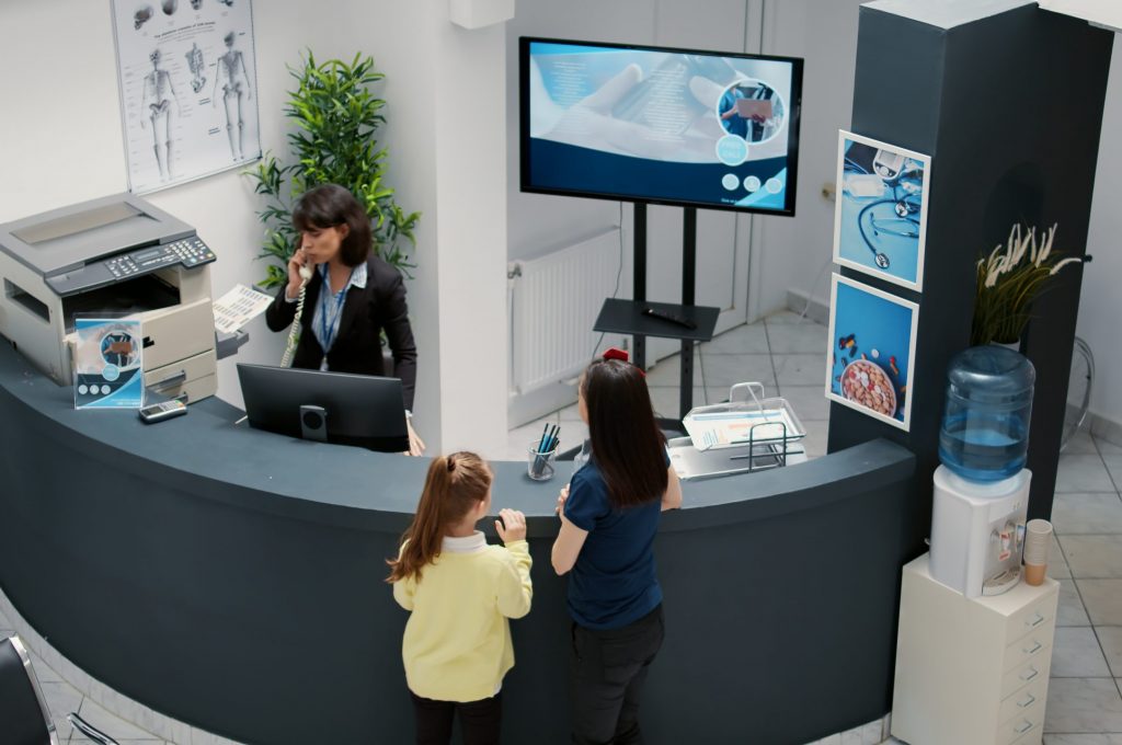 Receptionist working in hospital waiting area and offering support to patients at checkup visit, mother with child having medical appointment with physician. Reception desk at healthcare clinic.